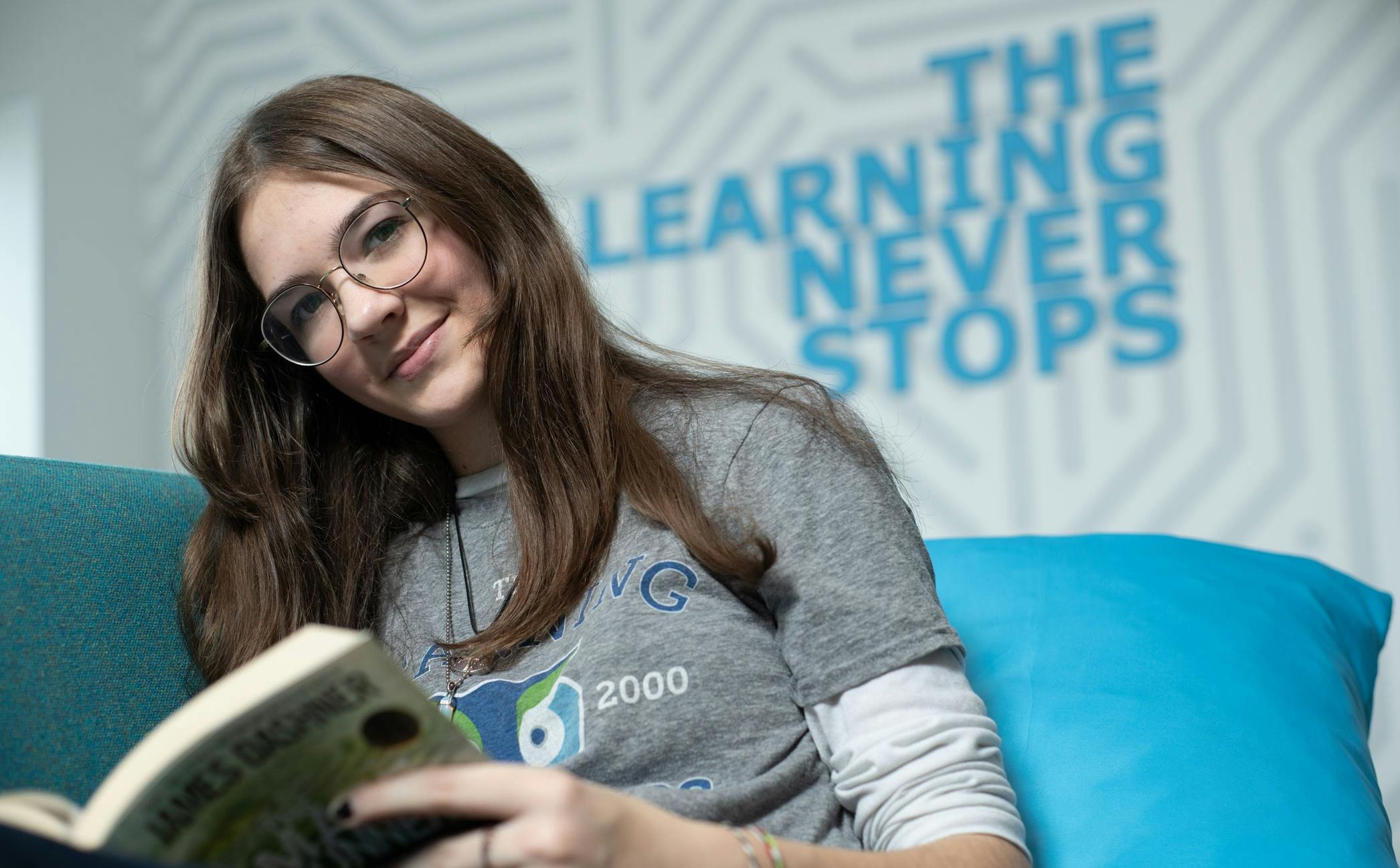 Teen student holds a book and smiles while looking at the camera. The rear wall says "The Learning Never Stops."