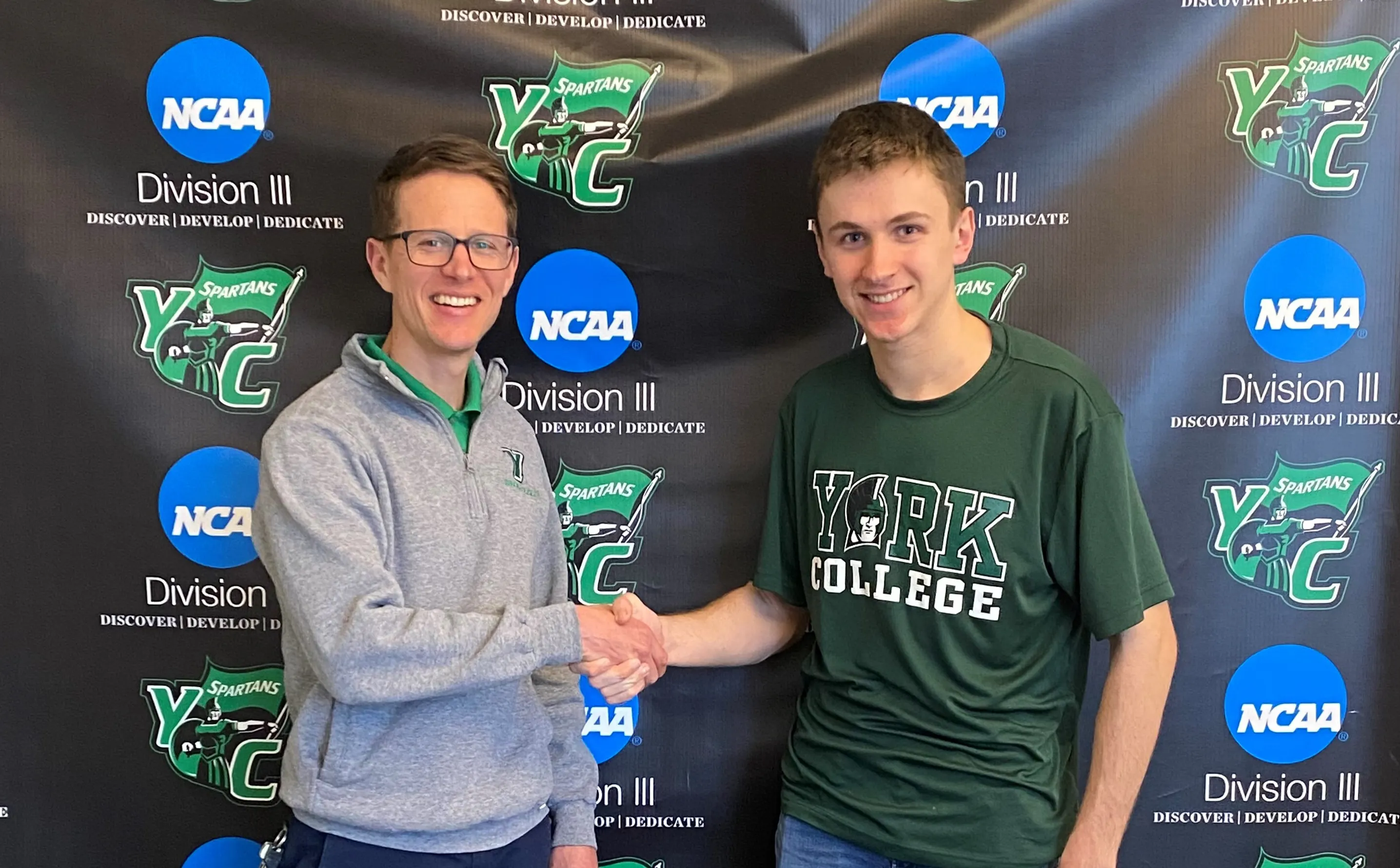 Student shakes hands with his future coach. They are standing in front of a branded York College backdrop.