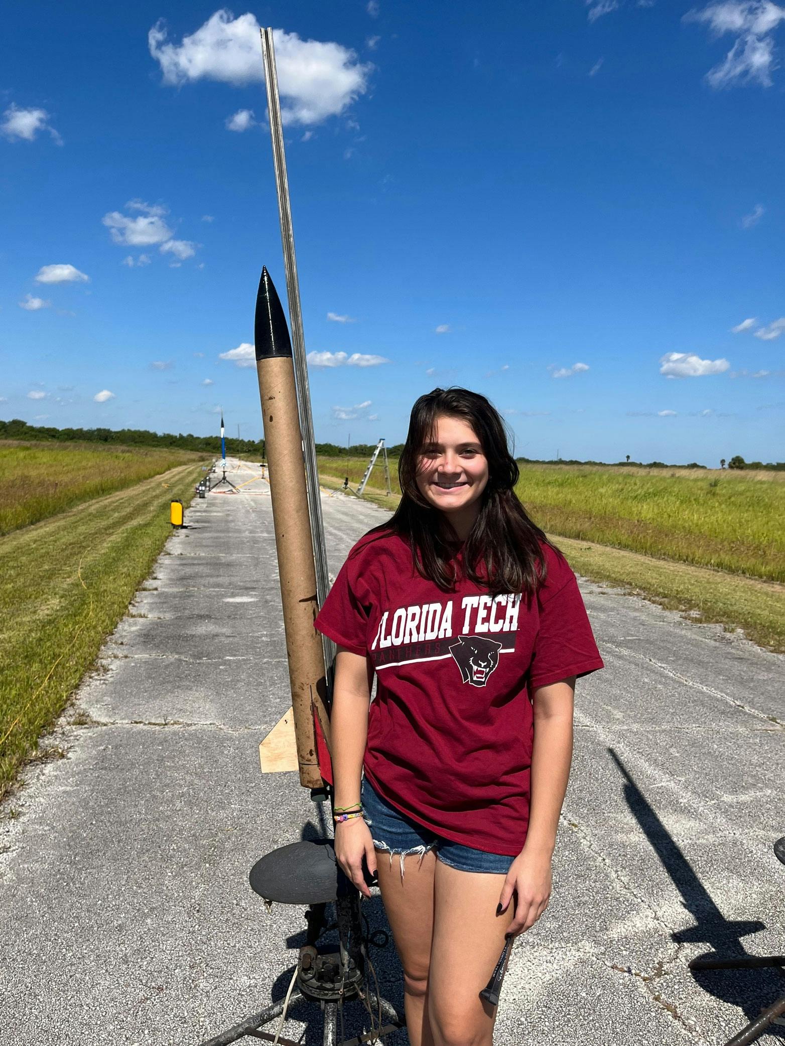 Karissa is standing in front of her rocket. She is wearing a Florida Tech T-shirt.