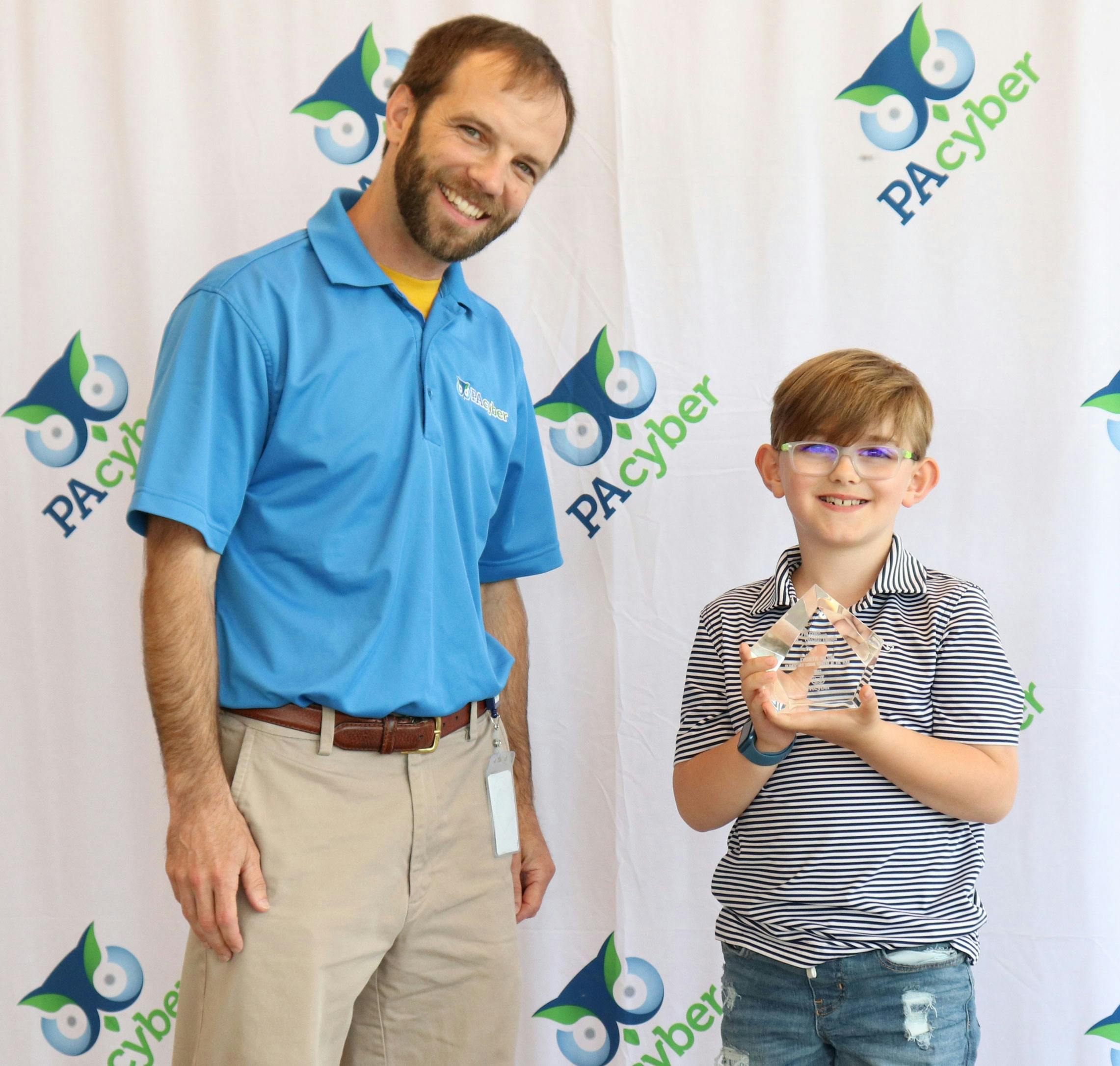 Young boy holds a pentagon-shaped glass award in front of a backdrop with the PA Cyber logo. A man with a PA Cyber-branded shirt stands next to him.